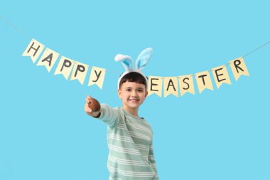 Tavşan kulaklı şirin bir çocuk elinde çikolata yumurtası ve mavi arka planda Mutlu Paskalya tatili yazılı kağıt bir çelenk tutuyor.