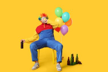 Balonlu ve bira şişeli alkolik palyaço gibi sarı arka planda oturan genç bir adam.