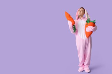 Paskalya tavşanı kostümlü düşünceli genç adam leylak arka planında peluş havuçlar tutuyor.