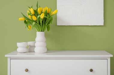 Çekmece dolabında sarı lale vazosu ve oturma odasındaki duvarda resim.