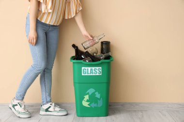 Bej duvarın yanında GLASS tabelası olan bir kadın şişeyi çöp kutusuna koyuyor. Geri dönüşüm kavramı