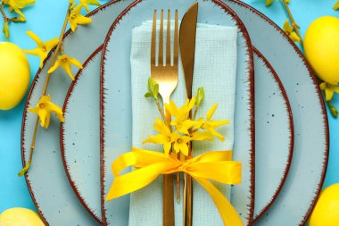 Mavi arka planda peçete, çatal bıçak takımı ve sarı çiçekli tabaklar.