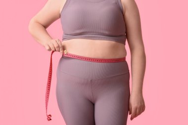 Şişman bir kadın, pembe arka planda belini ölçüyor. Diyet kavramı