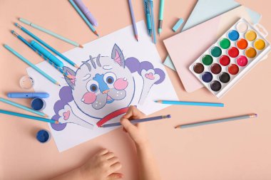 Kalemlerle, suluboya boyalarla ve bej arka planda farklı kırtasiyelerle çocuk eli çiziyor.