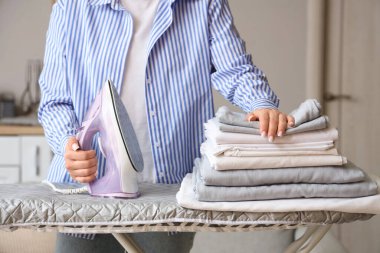 Katlanmış çarşaf ve ütü masasında ütü yığını olan bir kadın.