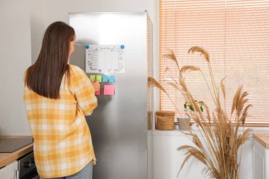 Modern mutfaktaki buzdolabına yemek planı yapıştıran genç bir kadın.