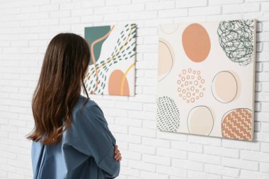 Sanat galerisinde beyaz tuğlalı duvardaki resimlere bakan genç bir kadın.