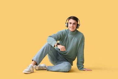 Sarı arka planda oturan, modern akıllı kol saati olan, şık gri kazaklı ve kulaklıklı yakışıklı genç adam.