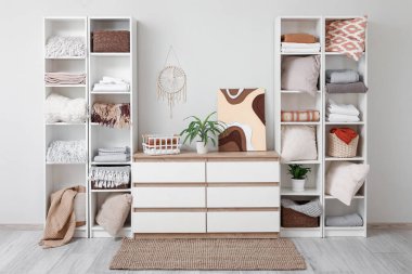 Odanın içindeki beyaz duvarın yanında temiz çarşaf, ekose ve sepetli raflar.