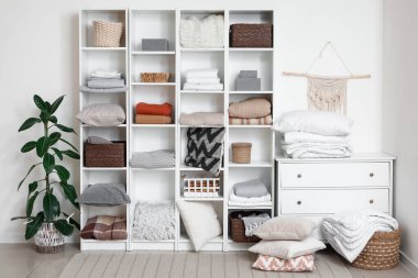 Odanın içinde beyaz duvarın yanında temiz çarşaflar, ekose ve sepetler olan raf ünitesi.