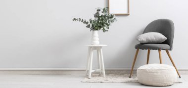 Odadaki beyaz duvarın yanındaki taburede okaliptüs dalları olan sandalye, puf ve vazo. Tasarım için pankart