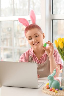 Paskalya yumurtası olan genç bir kadın mutfaktaki masada dizüstü bilgisayar kullanıyor.