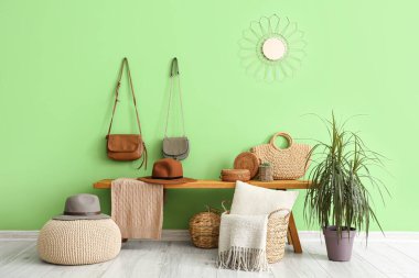 Hasır sepetli ahşap bank, yeşil duvarın yanında şapka ve çantalar.