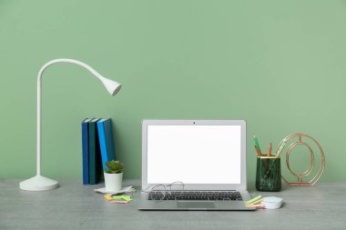 Yeşil duvarın yanında dizüstü bilgisayarı, kırtasiyesi ve lambası olan modern ofis çalışma alanı