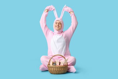 Paskalya tavşanı kostümü içinde oturan ve mavi arka planda kulakları olan mutlu genç adam.