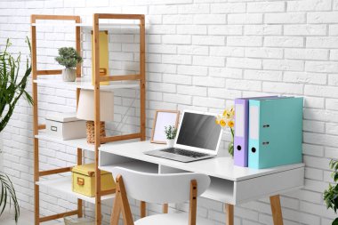 Ofisteki beyaz tuğla duvarın yanında bir buket narsisli çiçek ve dizüstü bilgisayarla raf ve işyeri.