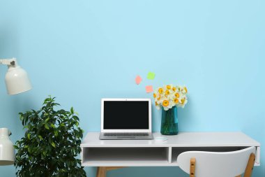 Şık bir ofiste mavi duvarın yanında bir buket narsisli çiçek ve dizüstü bilgisayarlı bir iş yeri.