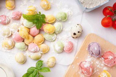 Baking paper and wooden board of colorful raw dumplings with basil on white background
