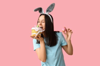 Tavşan kulaklı güzel bir kadın, pembe arka planda paskalya yumurtası ve pasta.