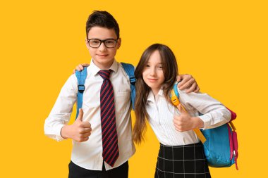 Sırt çantalı sevimli okul çocukları sarı arka planda baş parmak hareketi gösteriyor.