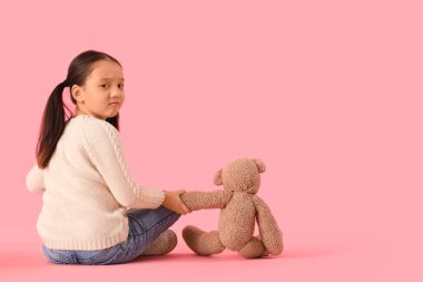 Otistik bozukluğu olan küçük bir Asyalı kız ve oyuncak ayı pembe arka planda oturuyor.