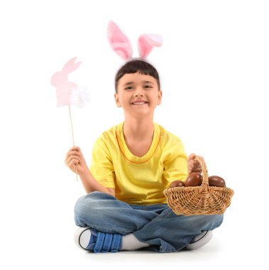 Beyaz arka planda Paskalya için çikolata yumurtası ve dekorla dolu sepet tutan tavşan kulaklı şirin bir çocuk.