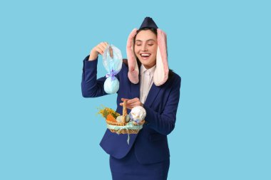 Tavşan kulaklı mutlu hostes, mavi arka planda çikolata yumurtası ve paskalya sepeti var.