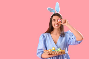 Tavşan kulaklı güzel bir kadın elinde hasır tepsiyle pembe arka planda Paskalya yumurtaları tutuyor.