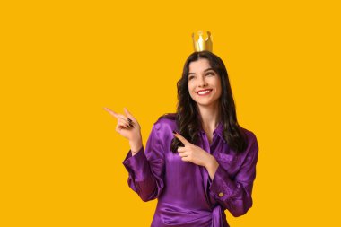 Şık balo elbisesi ve kağıttan taç giymiş güzel genç bir kadın sarı arka plandaki bir şeyi işaret ediyor.