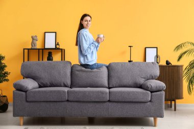 Genç bir kadın oturma odasında elinde fincanla kanepede oturuyor.