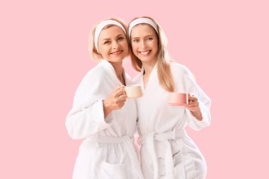 Mutlu genç kadın ve annesi pembe arka planda çay fincanlarıyla bornozlu.