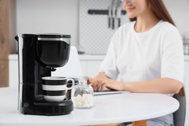 Modern kahve makinesi ve dizüstü bilgisayarı olan genç mutlu kadın mutfakta, yakın plan.