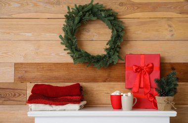 Şömine rafı, kışlık giysiler, hediye kutusu, sıcak çikolata ve ahşap duvarın yanında Noel çelengi.