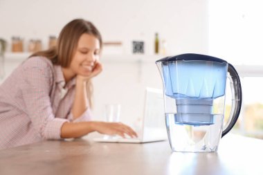 Mutfakta dizüstü bilgisayarı ve su filtresi olan genç bir kadın var.