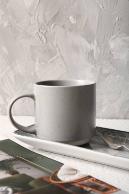 Gri duvarın yanındaki beyaz masada seramik bardaklar, tepsi ve dergi kompozisyonu