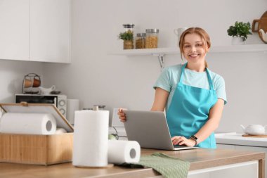 Kağıt peçeteli genç bir kadın mutfaktaki masada dizüstü bilgisayar kullanıyor.