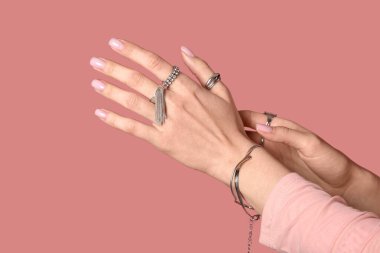 Pembe arka planda gümüş yüzüklü ve bilezik takan kadın elleri, yakın plan.