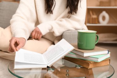 Elinde kahve ve kitaplarla evdeki kanepede oturan genç bir kadın.