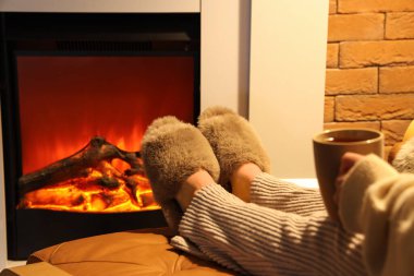 Terlikli bir kadın ve evinde şöminenin yanında oturan bir fincan çayla.