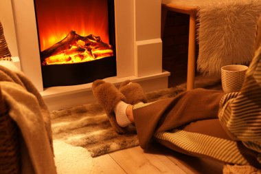 Terlikli bir kadın ve evinde şöminenin yanında oturan bir fincan çayla.