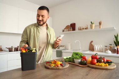 Mutfakta yemek pişirirken cep telefonu, gübre kutusu ve sebze artıkları olan genç bir adam.