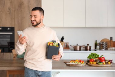 Cep telefonu, gübre kutusu ve sebze artıkları olan genç bir adam mutfakta.