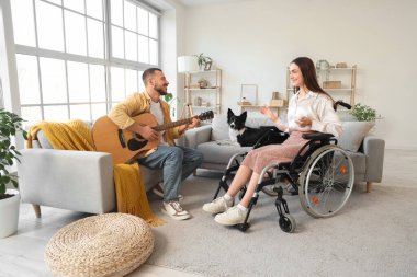 Tekerlekli sandalyede, kocası ve köpeği evde gitar çalan genç bir kadın.