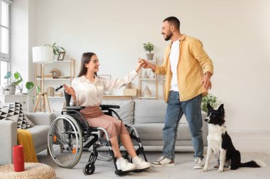 Tekerlekli sandalyedeki genç kadın ve evde dans eden kocası.