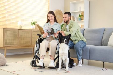 Tekerlekli sandalyedeki genç kadın, koca ve köpek okuma dergisiyle evde.