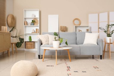 Oturma odasındaki bej duvarın yanında çiçekli gri kanepe ve boş poster.