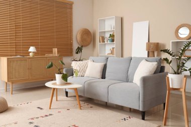 Oturma odasındaki bej duvarın yanında çiçekli gri kanepe ve boş poster.