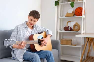 Genç çocuk evdeki kanepede gitar çalıyor.