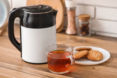 Modern elektrikli çaydanlık, mutfakta bir bardak sıcak çay ve tatlı kurabiyeler.
