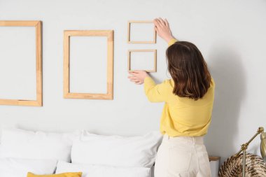 Genç bir kadın ışık duvarına ahşap çerçeveler asıyor. Yatak odası, arka manzara.